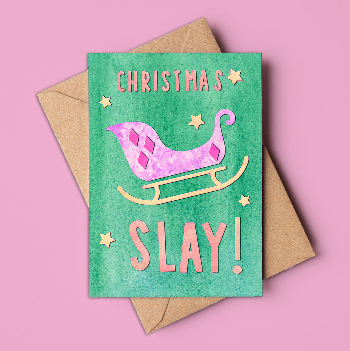A vibrant, non-traditional Christmas card made in Australia, featuring a pink and purple sleigh with gold accents on a green background. The card reads &quot;CHRISTMAS SLAY!&quot; in bold, playful lettering, surrounded by gold stars. Set against a pink backdrop with a kraft envelope, this A6 card is designed for modern holiday greetings and is available to wholesale buyers.