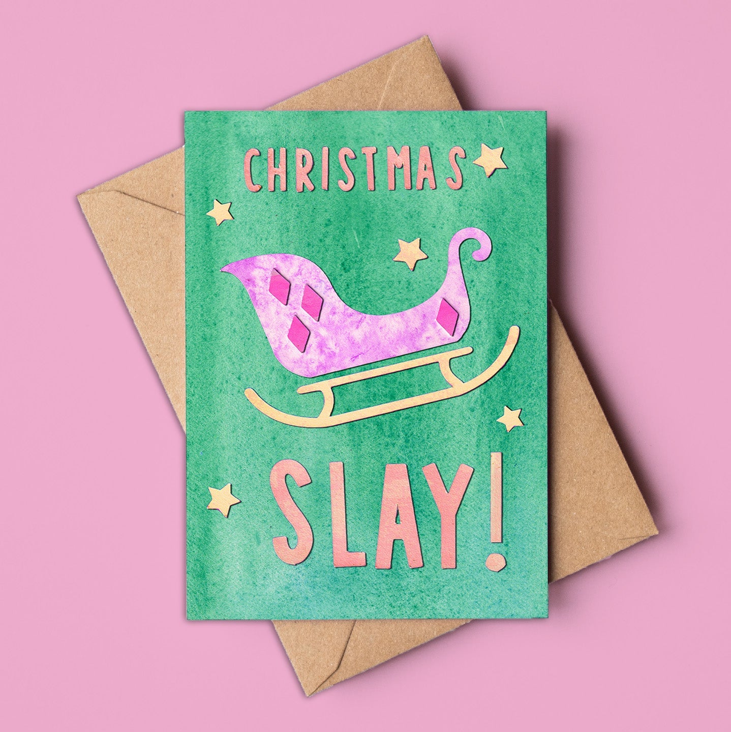 A vibrant, non-traditional Christmas card made in Australia, featuring a pink and purple sleigh with gold accents on a green background. The card reads "CHRISTMAS SLAY!" in bold, playful lettering, surrounded by gold stars. Set against a pink backdrop with a kraft envelope, this A6 card is designed for modern holiday greetings and is available to wholesale buyers.