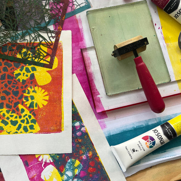 Colorful art supplies including a brayer, a gel plate, tubes of paint, stencils and printed artworks on a table.