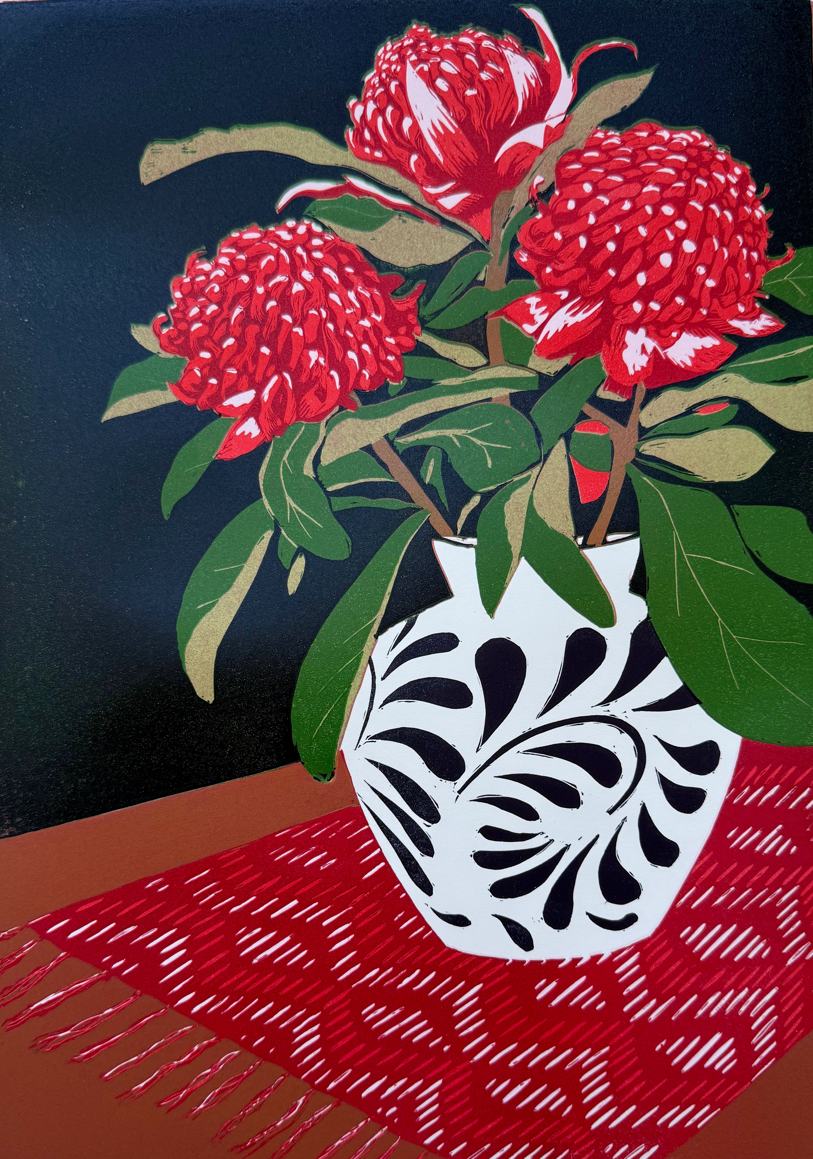A vase of red waratahs on a woven table runner on a wooden table, hand printed, reduction print
