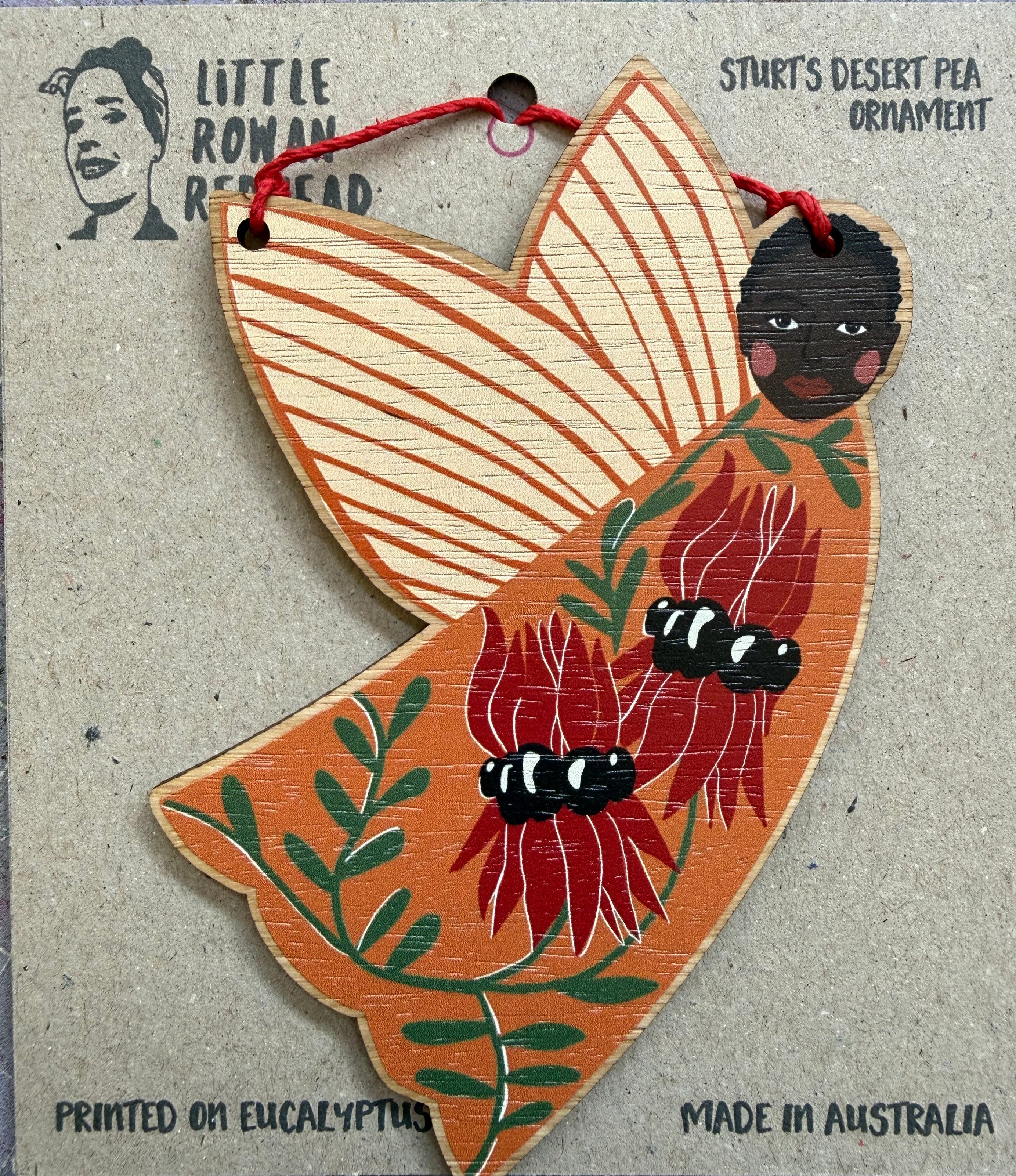 A wooden hanging fairy ornament decorated with red flowers and  green leaves. It is attached to a kraft paper backing.