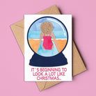 Card with a snowglobe illustration of a person in a swimsuit and text 'It's beginning to look a lot like Christmas' on a pink background. Made in Australia and available for wholesale.